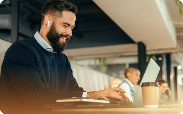Vīrietis ar baltām bezvadu austiņām strādā pie portatīvā datora kafejnīcā, uz galda atrodas kafijas krūze un piezīmju grāmatiņa, fonā ir citas personas.