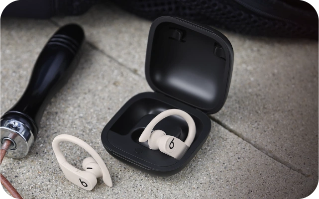 Divas bēšas bezvadu austiņas ar aizauss stiprinājumiem, viena novietota uz betona virsmas, otra ievietota melnā uzlādes futrālī.