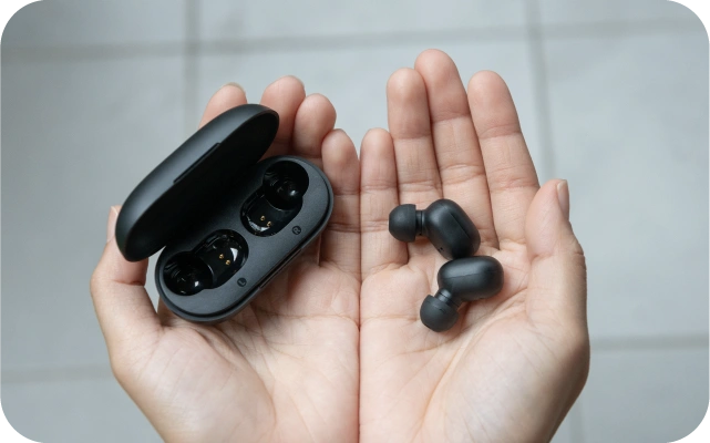 Cilvēka rokas tur divas melnas bezvadu austiņas un atvērtu uzlādes kārbiņu melnā krāsā.