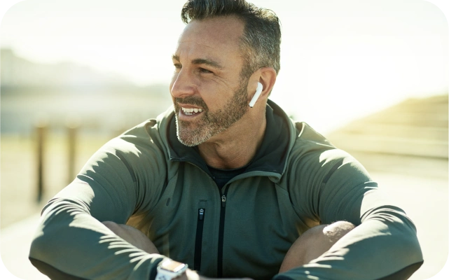 Vīrietis ar pelēku jaku un baltām bezvadu austiņām ausīs sēž ārā saulainā dienā.