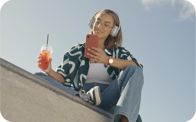 Meitene ar lielām austiņām ap galvu, zaļi baltu kreklu un dzērienu rokā sēž uz betona virsmas, skatoties mobilajā telefonā.