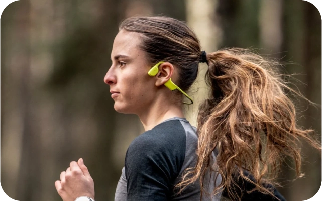 Sieviete ar gariem brūniem matiem, melnā sporta kreklā un dzeltenām open-ear austiņām skrien mežā.