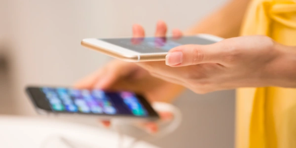 Persona dzeltenās drēbēs tur rokās divus mobilos telefonus; fokuss uz kreiso roku un iPhonu.
