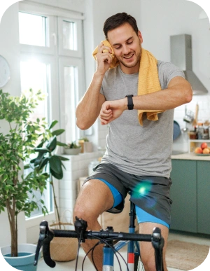 Vīrietis pelēkā T-kreklā un šortos uz velo trenažiera ar dzeltenu dvieli ap pleciem un skatās uz viedpulkstenī; fonā virtuve ar augiem un logu.