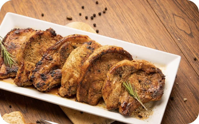 Balts šķīvis ar piecām cūkgaļas karbonādēm, kas dekorētas ar rozmarīnu; fonā koka galds un piparu graudiņi.