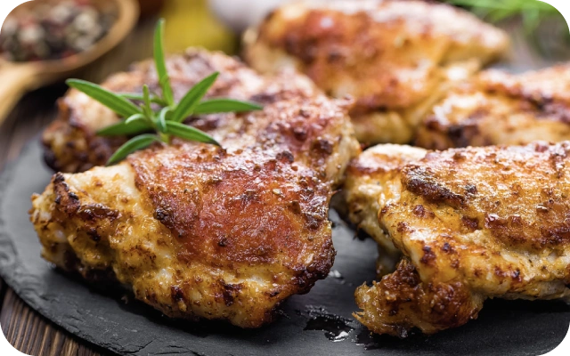 Zeltaini brūni cepti vistas šķiņķīši uz melna šķīvja, dekorēti ar svaigu rozmarīnu