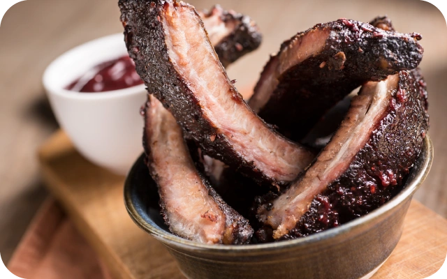 Bļoda ar sulīgām, glazētām grilētām cūkgaļas ribām, ar mērces trauciņu fonā.