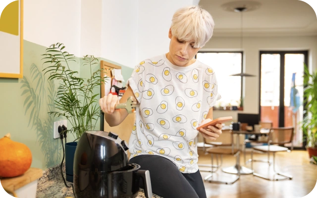 Sieviete darbojas virtuvē pie karstā gaisa fritera, turot rokā mobilo telefona un virtuves lāpstiņu; fonā zaļi augi un ēdamistaba.