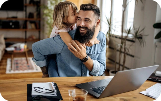 Vīrietis zilā kreklā sēž pie galda ar portatīvo datoru, piezīmju kladi, brillēm un tējas krūzi uz galda, kamēr bērns apskauj viņu no aizmugures.