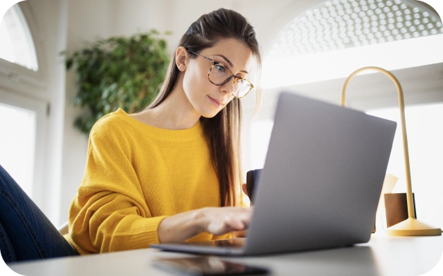 Sieviete ar gariem matiem, brillēm un dzeltenā džemperī strādā pie klēpjdatora, vienā rokā turot kafijas krūzi.
