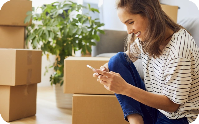 Sieviete svītrainā t-kreklā un zilās biksēs sēž uz grīdas un skatās mobilajā telefonā, apkārt vairākas kartona kastes zaļš istabas augs.