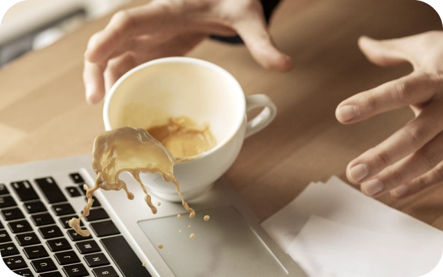 Cilvēks nejauši izlej kafiju uz portatīvā datora tastatūras, mēģinot to apturēt ar rokām; uz galda papīri ar grafikiem.