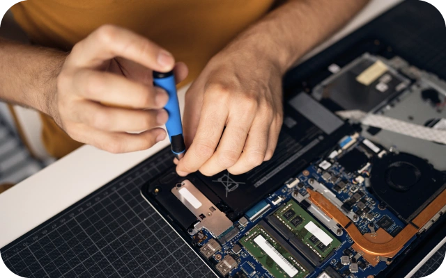Vīrietis rokās tur skrūvgriezi veic remontdarbus portatīvajam datoram, kas stāv uz galda; redzama mātesplate un atmiņas moduļi.