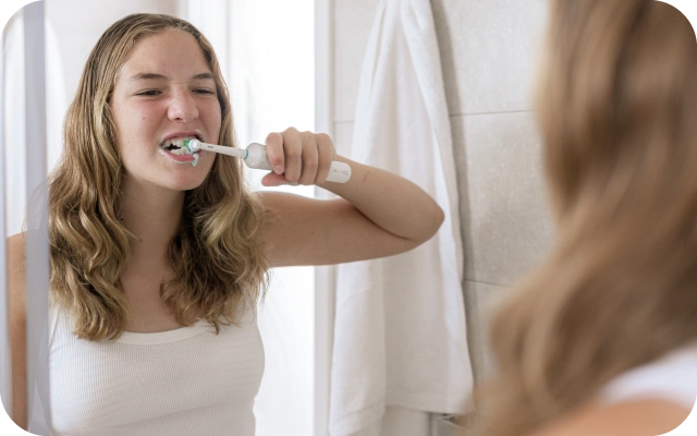 Meitene ar gariem viļņainiem matiem, baltā krekliņā, tīra zobus ar elektrisko zobu birsti vannas istabā.