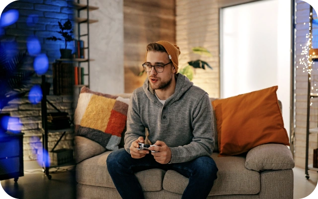 Vīrietis pelēkā džemperī un cepurē sēž uz dīvāna un spēlē videospēli ar kontrolieri; fonā krāsaini spilveni un plaukts mājīgā telpā.