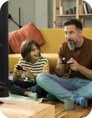 Vīrietis un bērns sēž uz grīdas dzīvojamā istabā un spēlē videospēles ar kontrolieriem, fonā dzeltens dīvāns.