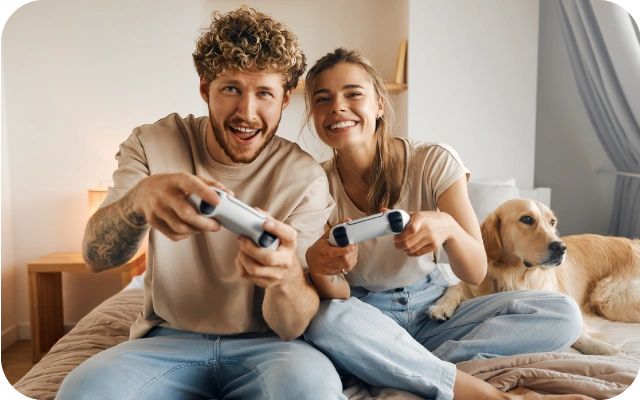 Sieviete un vīrietis sēž uz gultas un spēlē videospēles ar kontrolieriem, blakus gultā guļ liels suns.