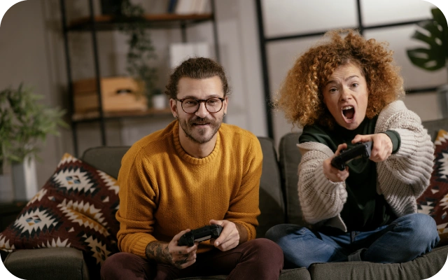 Vīrietis un sievete sēž uz dīvāna un spēlē videospēles ar kontrolieriem; vīrietis valkā dzeltenu džemperi, sievietei gaiša adīta jaka un zilas bikses. Fonā plaukti un istabas augi.