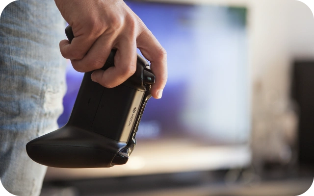 Cilvēks tur rokā Xbox spēļu kontrolieri, fonā izplūdis televizora ekrāns.
