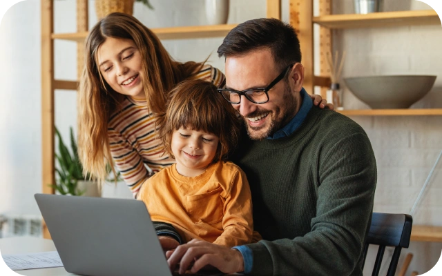 Ģimene pie galda skatās uz portatīvo datoru – vīrietis raksta, zēns sēž viņam klēpī, meitene stāv blakus, fonā plauktu sistēma ar dažādiem piederumiem.