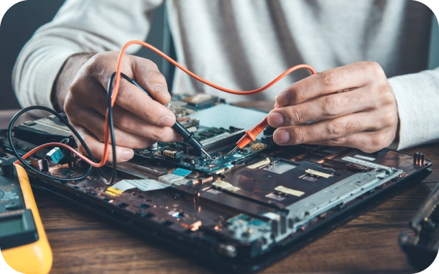 Tehniķis pelēkā džemperī pārbauda portatīvā datora mātesplati ar multimetru, fokuss uz portatīvā datora mātesplati.