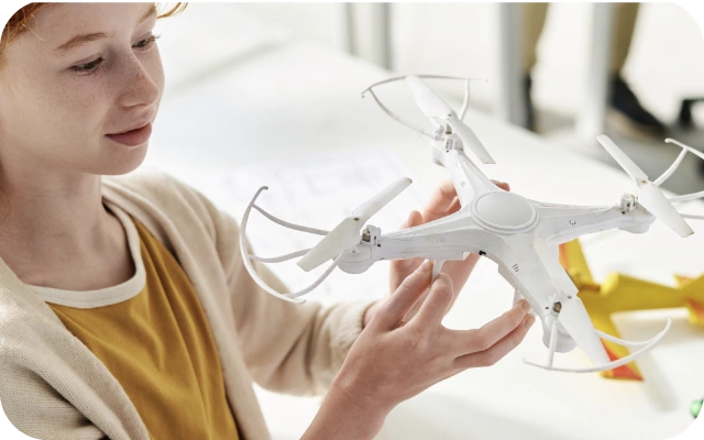 Bērns dzeltenā kreklā un gaišā jakā tur rokās baltu dronu.