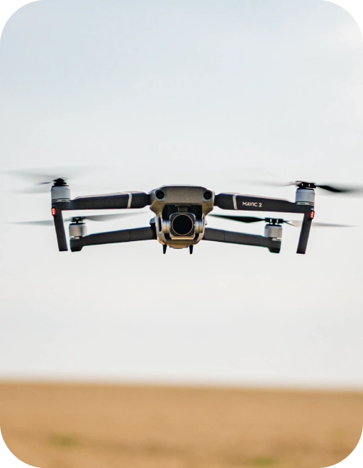 Pelēks Mavic 2 drons ar kameru lido gaisā virs lauka.