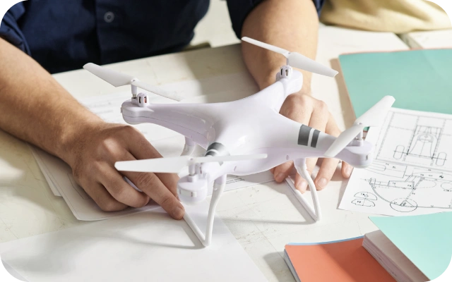 Cilvēks sēž pie galda un tur baltu dronu, kas stāv uz galda; fonā ir dokumenti un zīmējumi, kas saistīti ar drona dizainu.