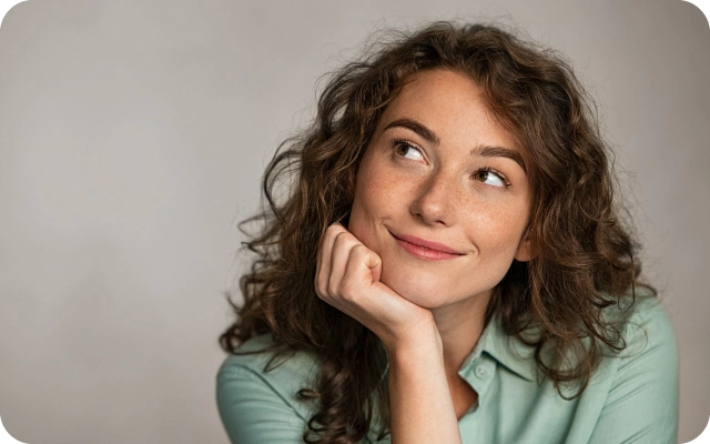 Sieviete ar cirtainiem matiem un gaiši zaļu kreklu, balsta galvu uz rokas.