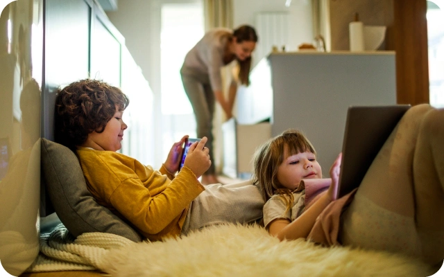 Divi bērni atpūšas uz grīdas noklāta pūkainas segas – viens spēlē ar mobilo telefonu, otrs spēlē planšetdatoru; fonā pie virtuves letes ir sieviete.