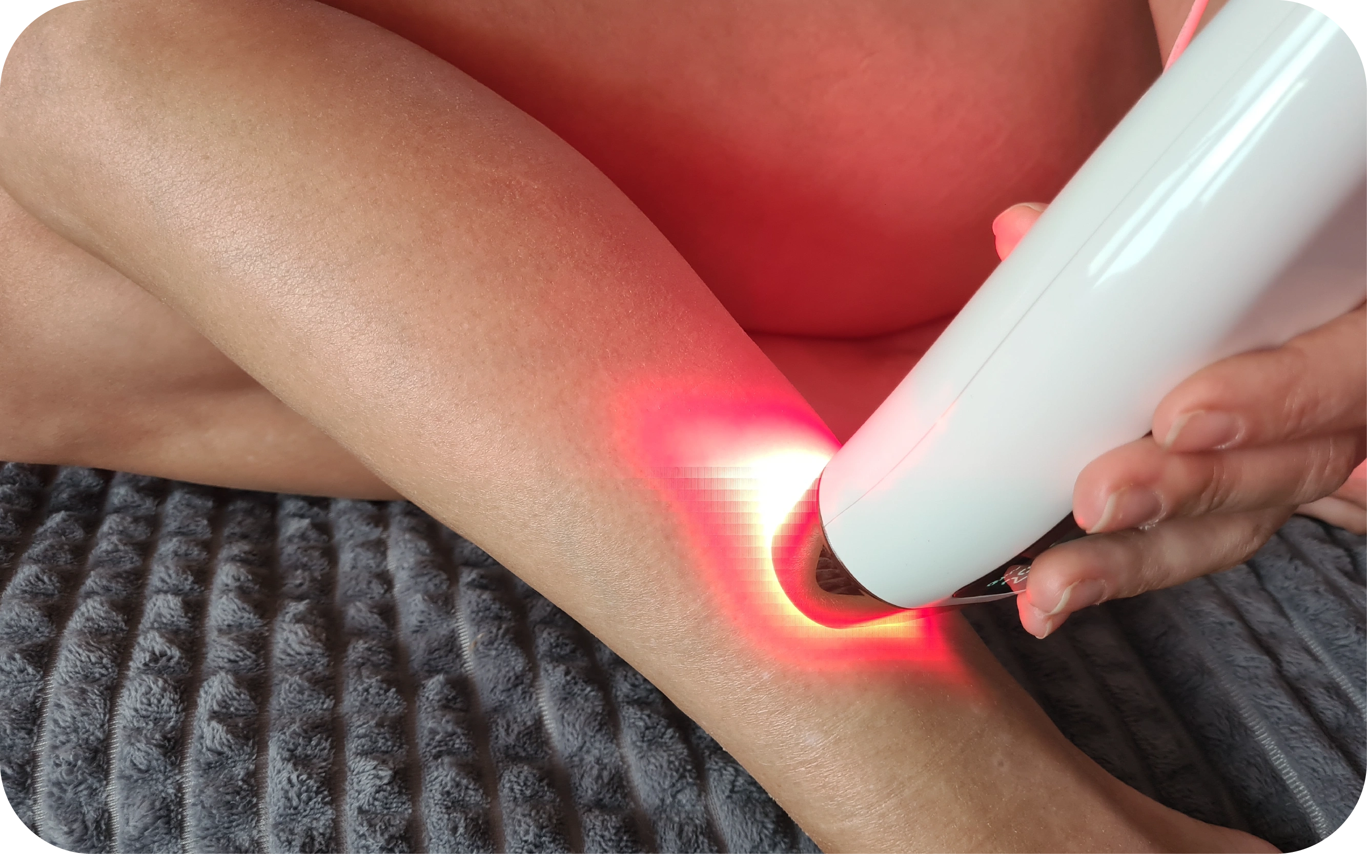 Persona izmanto baltu rokas lāzera epilatoru un epilē matiņus uz kājas; ierīce izstaro sarkanu gaismu.