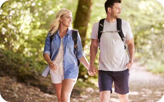 Vīrietis un sieviete ar mugursomām uz pleciem un sadevušies rokās pastaigājas pa saulainu meža taku.