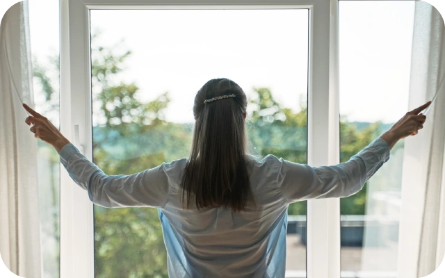 Sieviete pie liela loga atver aizkarus, aiz kura ir zaļi koki un gaišas debesis.