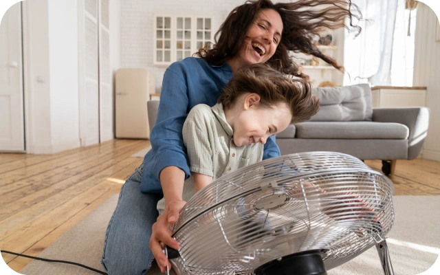 Sieviete un bērns sēž uz grīdas mājīgā viesistabā; sieviete tur rokās lielu gaisa ventilatoru, kas pūš vēso gaisu virsū.