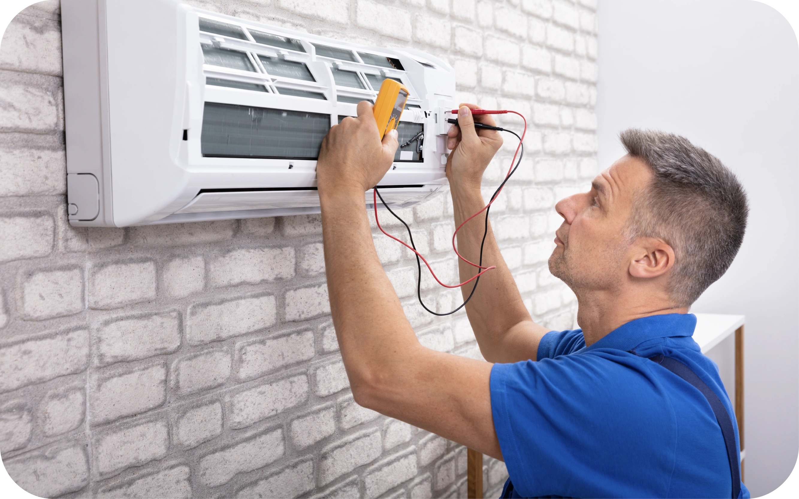 Vīrietis zilā kreklā ar multimetru pārbauda gaisa kondicionieri, kas piestiprināts pie baltas ķieģeļu sienas.