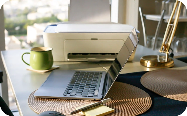 Darba galds ar atvērtu portatīvo datoru, zaļu krūzi, zelta galda lampu un printeri fonā. Uz galda ir apaļi galda paliktņi un piezīmju lapiņa ar pildspalvu.