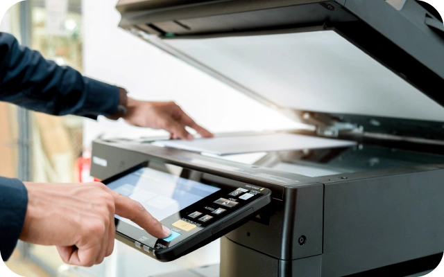 Tuvplāns ar personu, kas izmanto multifunkcionālo printeri ar skenēšanas iekārtu. Viena roka tur dokumentu uz stikla virsmas, otra pieskaras vadības panelim ar ekrānu un pogām.