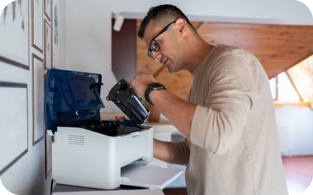 Vīrietis stāv pie printera ar atvērtu vāku, ievieto vai izņem tonera kasetni. Printeris ir balts ar zilu caurspīdīgu vāku, kas novietots uz galda telpā.