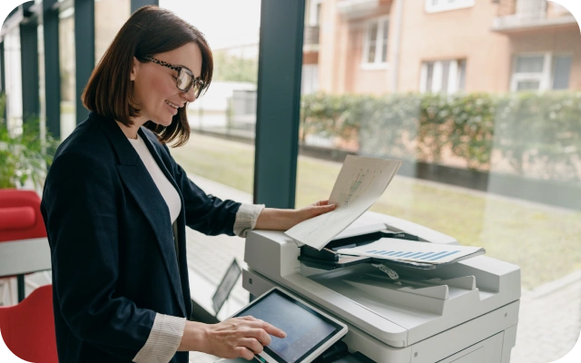 Sieviete tumšā žaketē stāv pie multifunkcionālā printera biroja vidē, tur dokumentu virs ierīces un ar otru roku pieskaras vadības panelim.