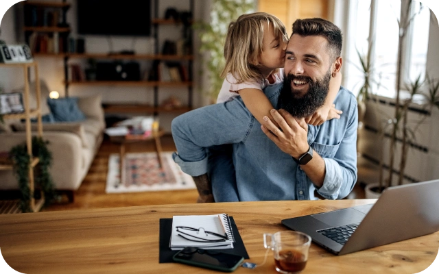 Vīrietis zilā kreklā sēž pie galda ar portatīvo datoru, piezīmju kladi, brillēm un tējas krūzi uz galda, kamēr bērns apskauj viņu no aizmugures.