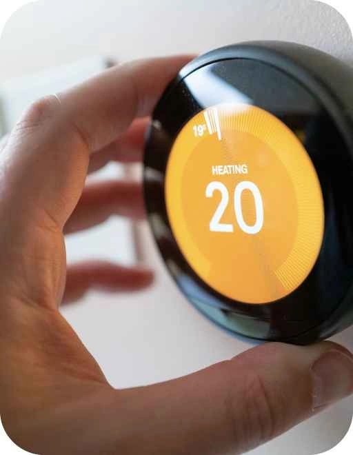 Cilvēka roka regulē melnu apaļu termostatu ar oranžu ekrānu, kurā rakstīts 'HEATING' un temperatūra 20 °C.