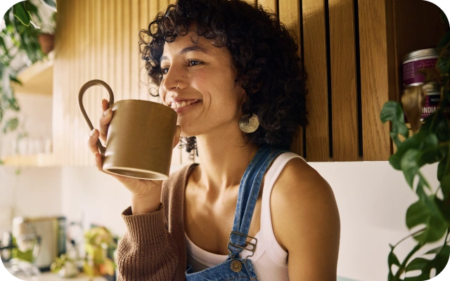 Sieviete ar cirtainiem matiem džinsa kombinezonā un gaišā topiņā stāv virtuvē un dzer no brūnas krūzes.