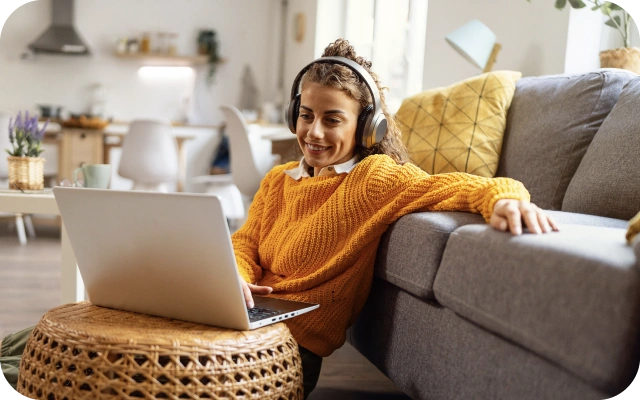 Sieviete ar austiņām sēž uz grīdas pie dīvāna un strādā ar portatīvo datoru, kas nolikts uz pīta galdiņa.
