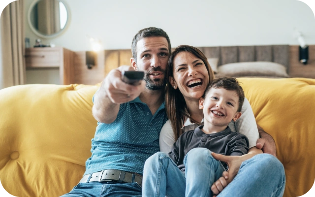 Vīrietis, sieviete un bērns kopā sakļāvušies sēž uz dzeltena dīvāna un skatās televizoru; vīrietis rokā tur pulti.