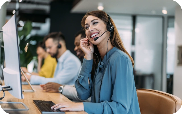 Sieviete zilā blūzē sēž pie darba galda un strādā pie datora ar austiņām un mikrofonu, fonā cilvēki, kas dara līdzīgu darbu.