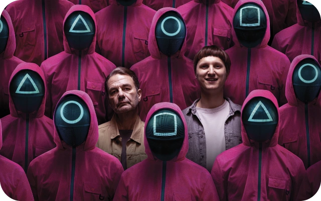 Divi vīrieši ikdienišķā apģērbā pa vidu cilvēku grupai rozā kombinezonos ar kapucēm un melnām maskām, uz kurām ir simboli – trijstūri, apļi un kvadrāti.