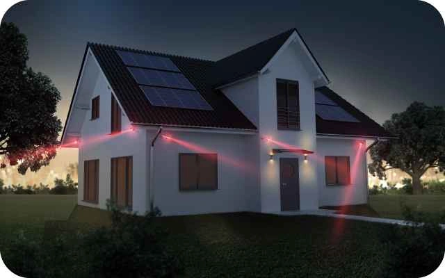 Balta māja ar saules paneļiem uz jumta un sarkanām gaismām no video novērošanas kamerām, fons ir tumšs ar dažiem kokiem apkārt un nelielu apgaismojumu aiz mājas.