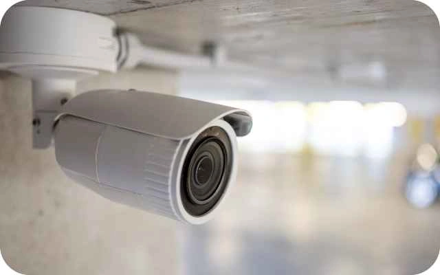 Video novērošanas kamera baltā krāsā uzstādīta pie griestiem, kas vērsta uz priekšu telpas novērošanai.