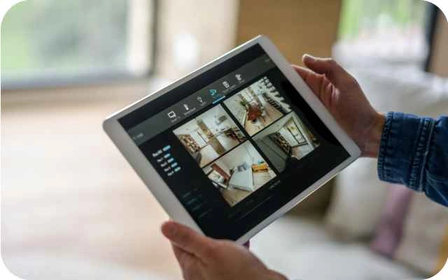 Persona tur rokās planšetdatoru ar atvērtu mājas drošības kameru lietotni, kurā ir četri dažādi skati no mājokļa iekšpuses.