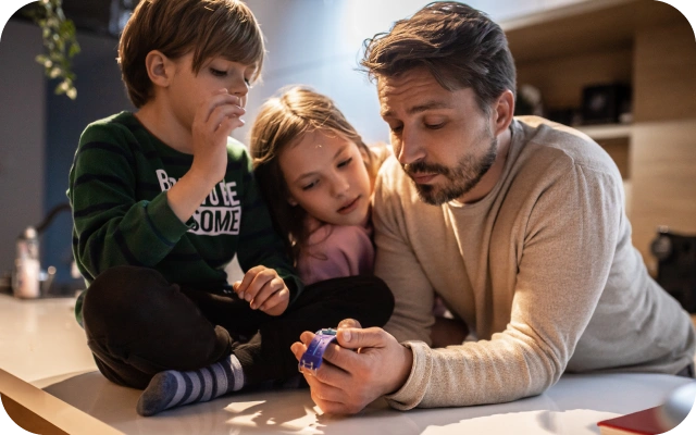 Vīrietis un divi bērni atrodas virtuvē pie galda virsmas un aplūko bērnu viedpulksteni ar violetu siksniņu, ko pieaugušais tur rokās. Priekšplānā viedpulkstenis, bet fonā plaukti, virtuves zona un silts iekštelpu apgaismojums.